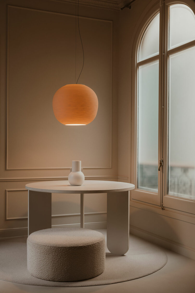 Minimalist room with a round table, white vase, textured ottoman, large window, and a hanging round pendant lamp emitting warm light.