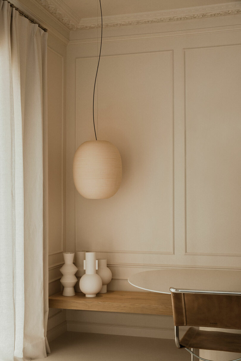 Minimalist beige room with a round hanging light, sheer curtains, a wooden bench with white ceramic vases, a round glass table, and a brown chair.