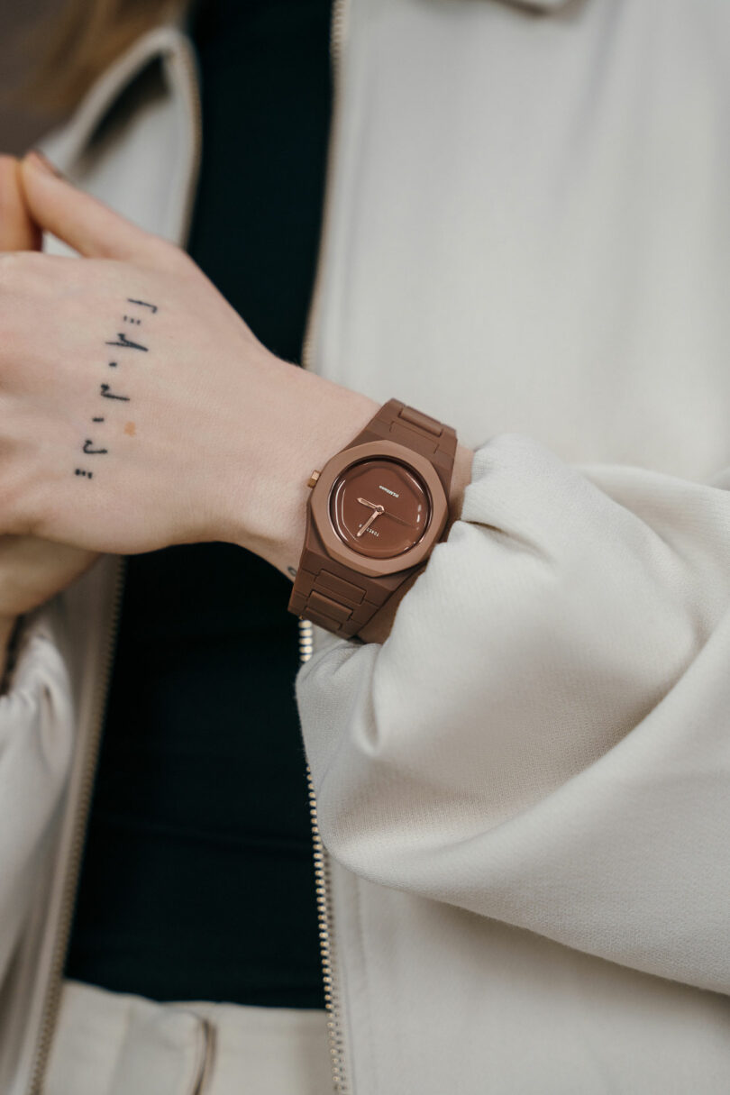 En la foto, una persona lleva un reloj marrón, una chaqueta ligera y un pequeño tatuaje negro en la mano.