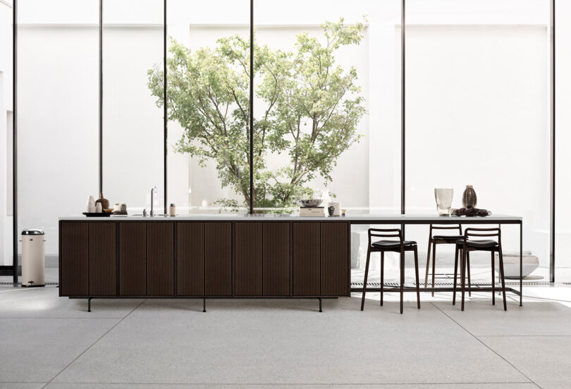 La moderna cocina cuenta con una isla Vipp de madera oscura, una decoración minimalista, tres taburetes y una gran ventana con un árbol verde afuera.