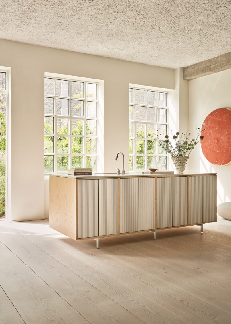 La cocina minimalista Vipp cuenta con una isla central, grandes ventanales, pisos de madera clara, jarrones y llamativos cuadros circulares rojos en las paredes.