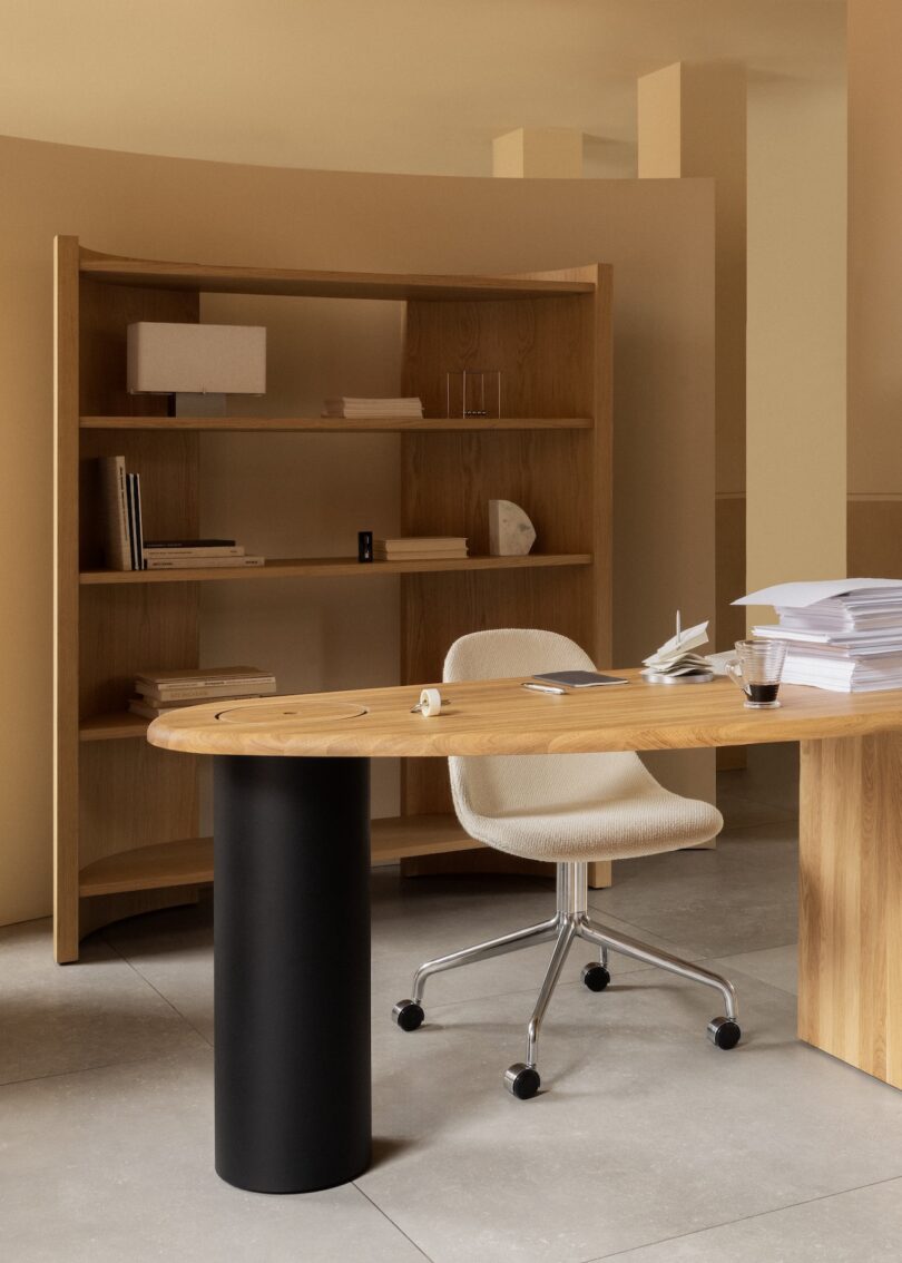 Una oficina moderna en una habitación de tonos neutros con un escritorio de madera, una silla con ruedas, una pila de papeles, una taza de café y una estantería vacía.