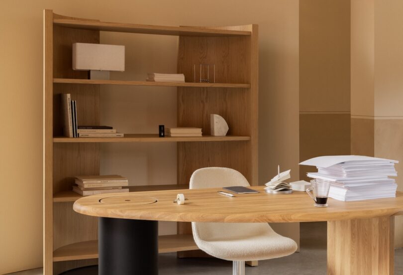 Oficina moderna con escritorio de madera, sillas color crema, documentos apilados, cuadernos, bolígrafos, tazas de café y estanterías de madera para libros y adornos.