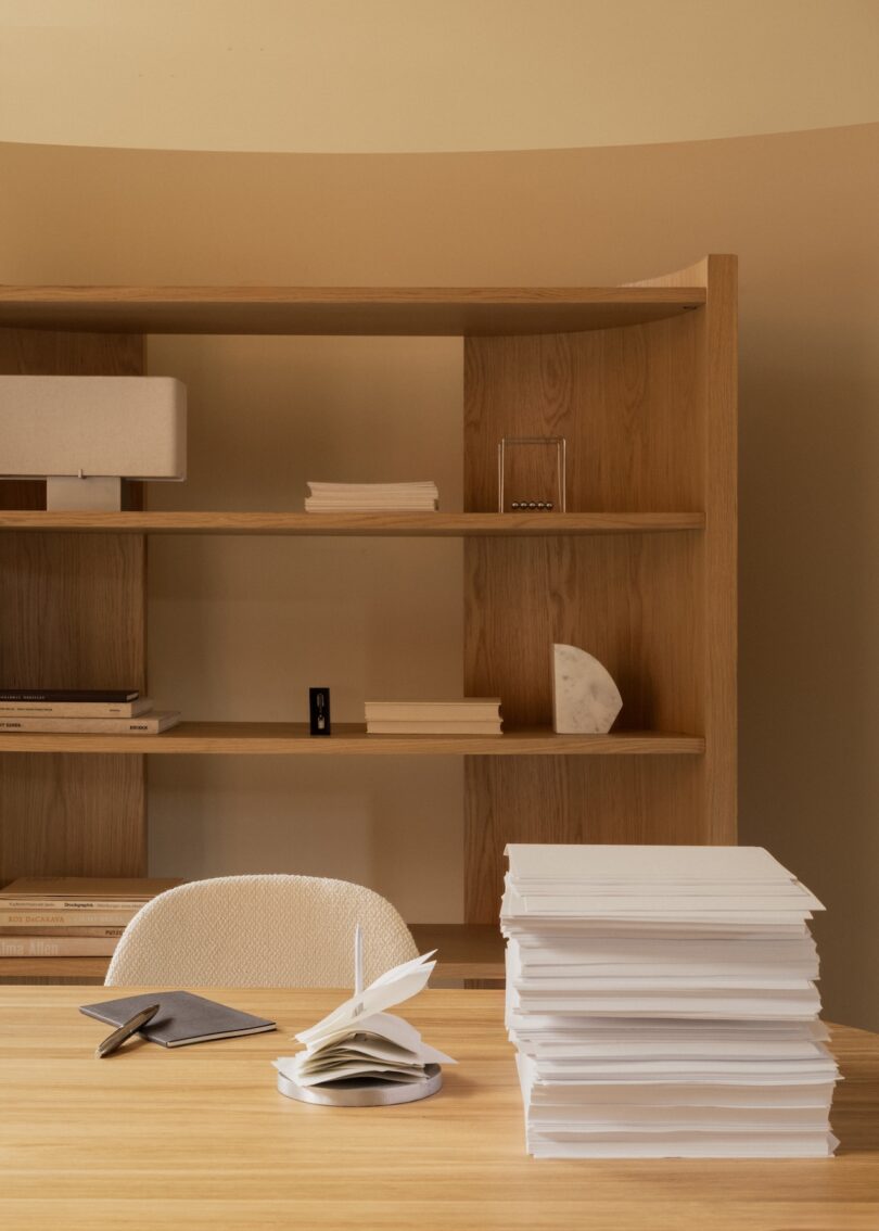 Una mesa de madera con una pila de papeles, un cuaderno abierto y un bolígrafo frente a una silla vacía; estantería de madera con libros y objetos al fondo.