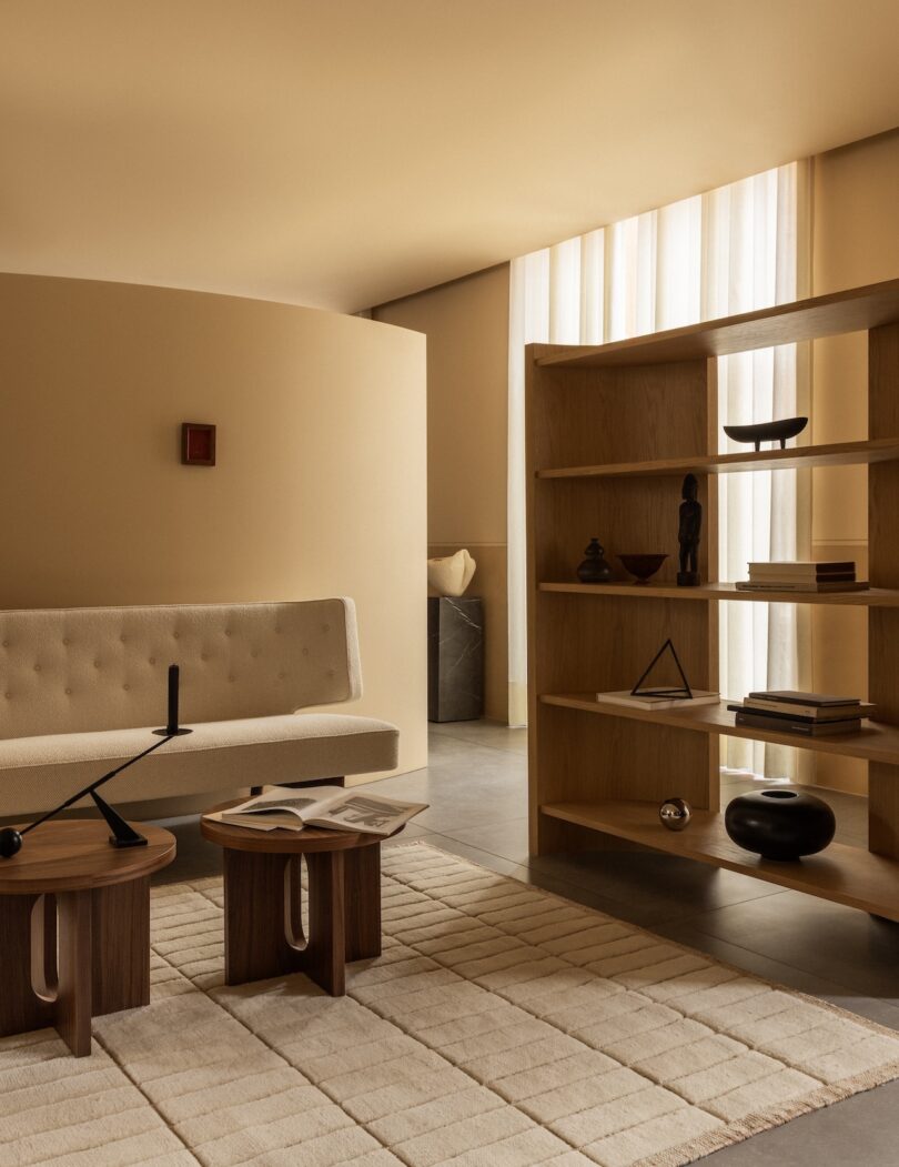 Salón moderno con paredes beige, sofá color crema, mesa de madera, alfombra clara y estantes abiertos de madera con libros y adornos. La luz natural entra a raudales a través de las altas cortinas.