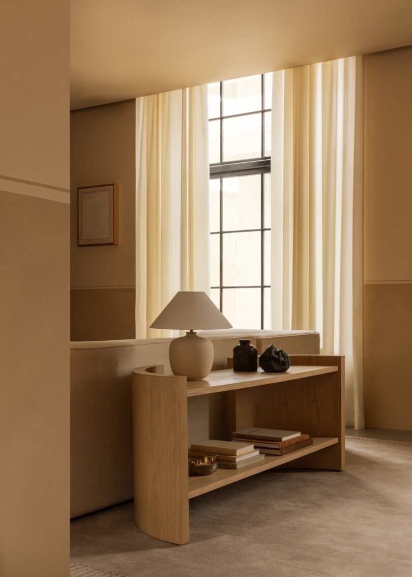 Salón sencillo con cortinas transparentes, ventanas, consola de madera clara con libros, una lámpara y elementos decorativos detrás de un sofá beige.