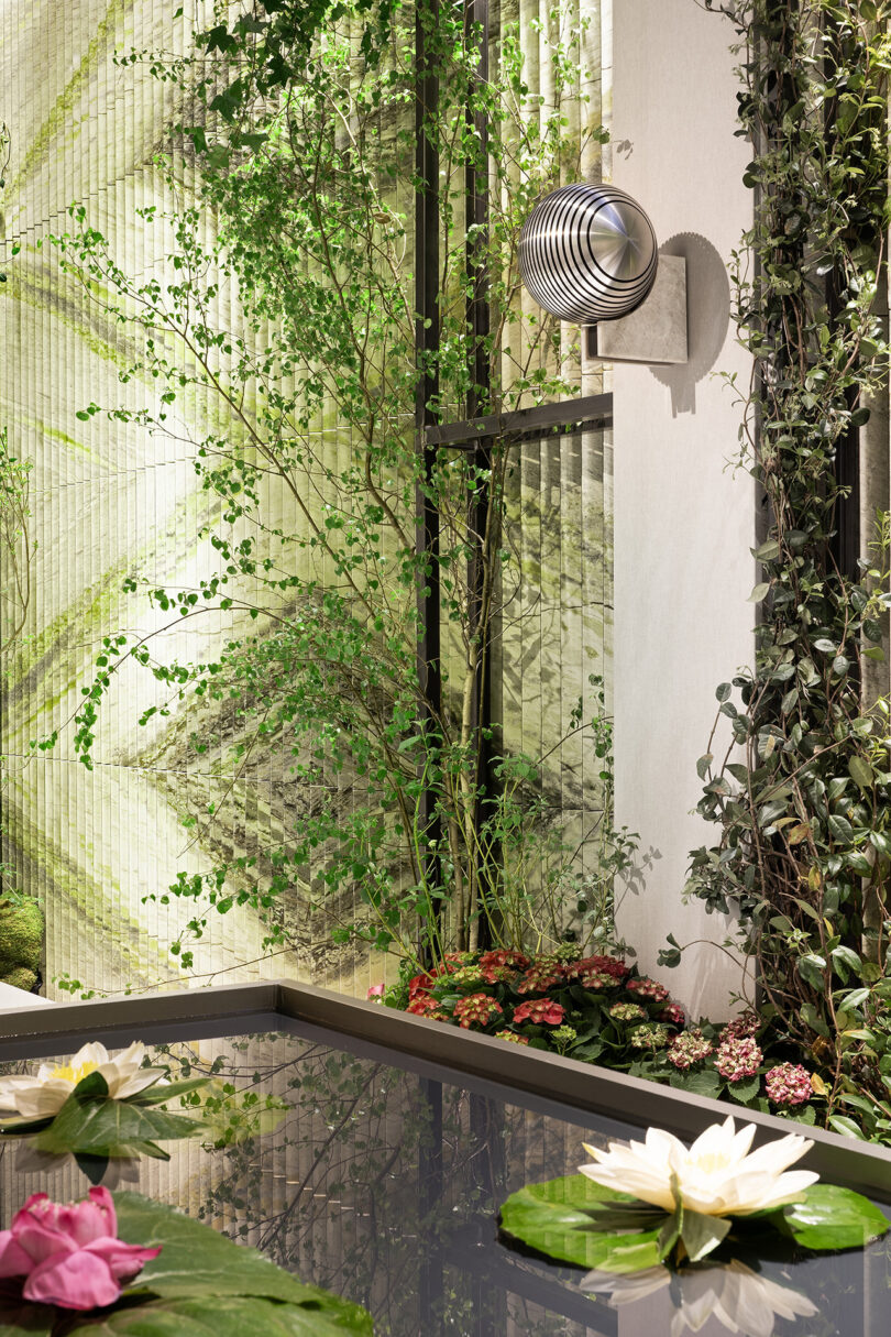Indoor garden scene with a pond featuring water lilies, green plants climbing the walls, and a modern metallic wall-mounted fan above the water.