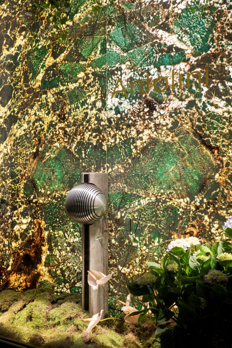A metallic sculpture with stacked spheres stands on moss beside flowers, set against a marbled green and gold backdrop with the word "Antolini" visible.