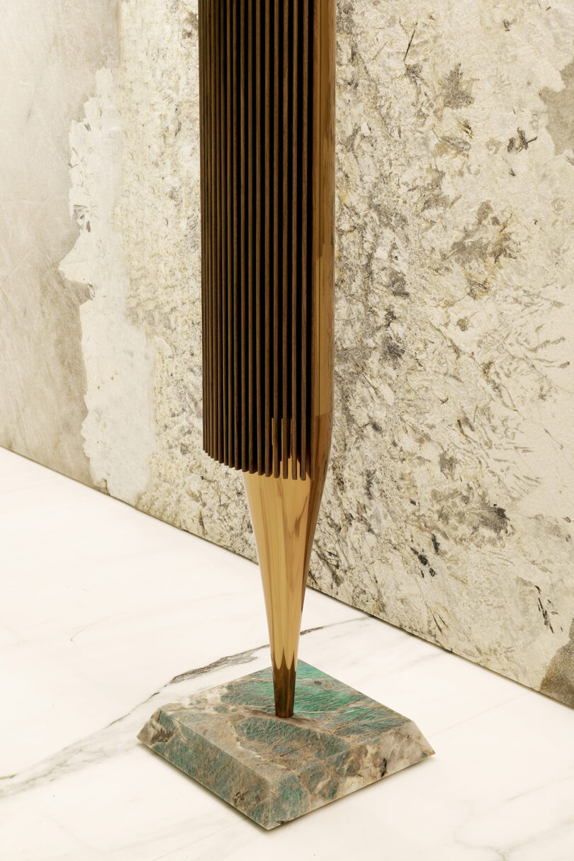 A tall, vertical metallic sculpture with ribbed sides tapers to a point and stands on a square greenish stone base, set against a textured stone wall.