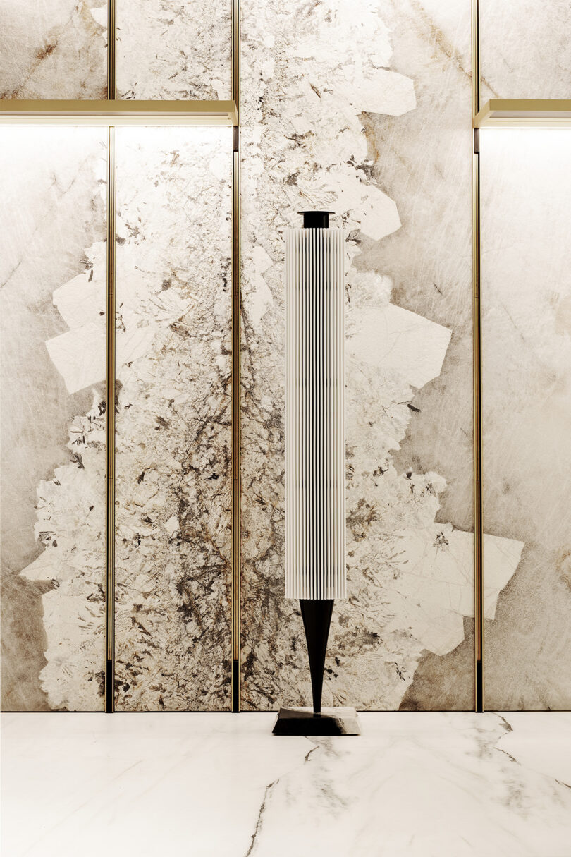 A tall, slim speaker with vertical white slats and a black base stands in front of a marble-patterned wall with gold accents on a marble floor.