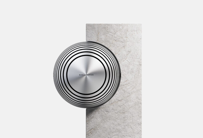 Round, metallic speaker with concentric black circles mounted on a vertical slab of light-colored stone, set against a plain white background.