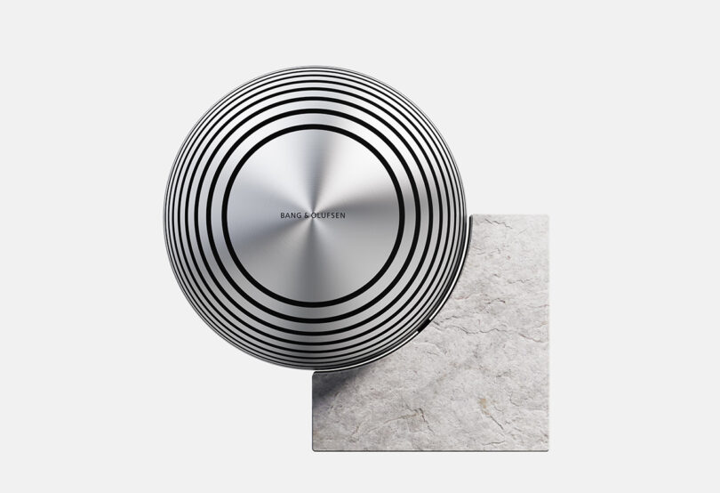 A round, silver Bang & Olufsen speaker with black concentric circles rests on a square piece of textured light gray stone against a plain white background.