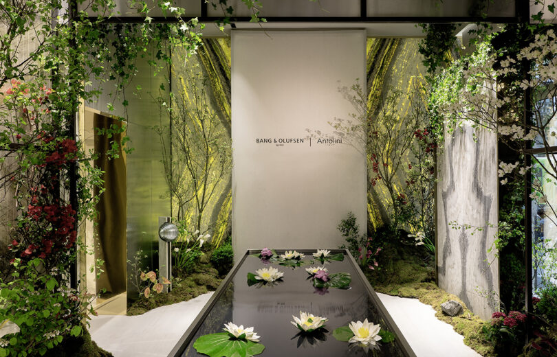 A modern indoor display featuring a reflective water surface with floating lotus flowers, surrounded by greenery, flowers, and a Bang & Olufsen and Annabel’s logo on the wall.
