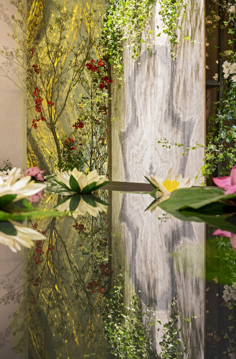 Water lilies float on a reflective pond, surrounded by greenery and flowers, with painted murals of trees and leaves in the background.