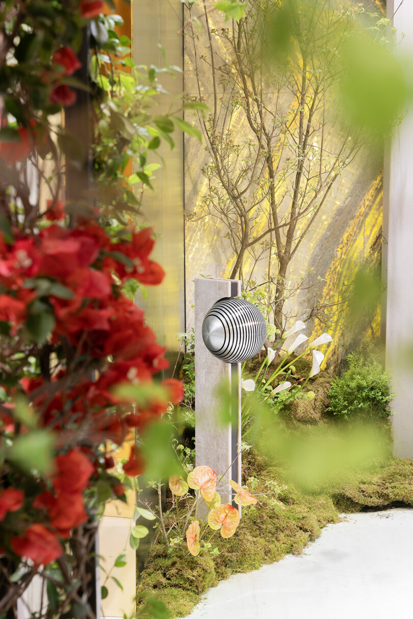 Modern outdoor speaker mounted on a post among green plants and red flowers in a landscaped garden setting.