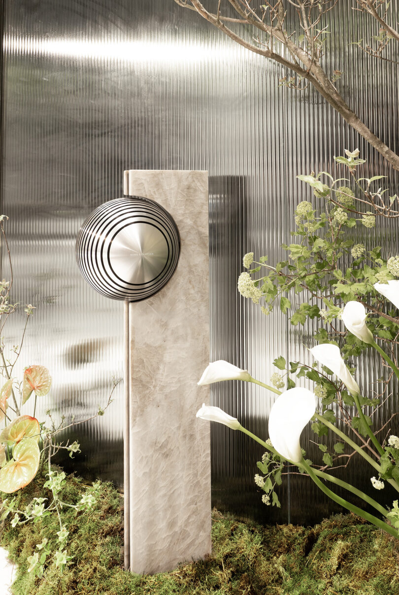 A modern sculpture with concentric metal circles on a rectangular marble base stands among green plants and white flowers against a metallic ribbed background.