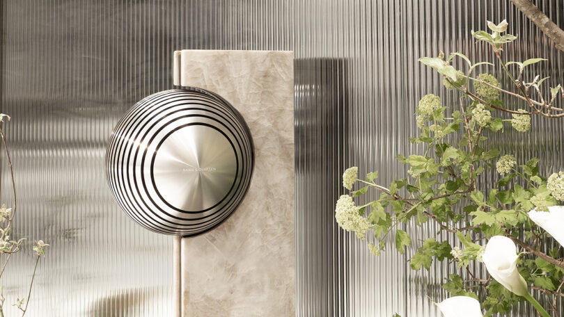 A circular metallic speaker with concentric rings is mounted on a marble stand, placed beside glass panels and flowering plants.
