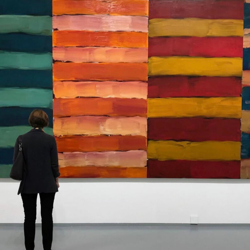 A person stands in front of a large abstract painting by Barry Ginder, featuring horizontal stripes in green, orange, red, and yellow tones in an art gallery.