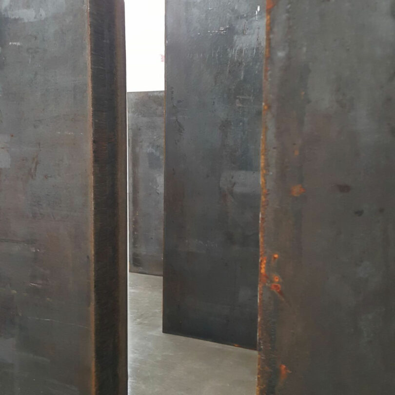 Large vertical metal slabs by Barry Ginder are arranged in parallel rows, creating narrow pathways on a concrete floor in an indoor space.