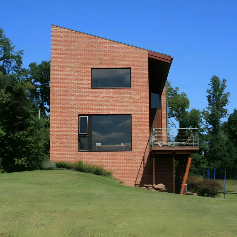 A modern two-story house with a slanted brick exterior, large black-framed windows, and a small balcony designed by Barry Ginder, set on a grassy slope with trees in the background.