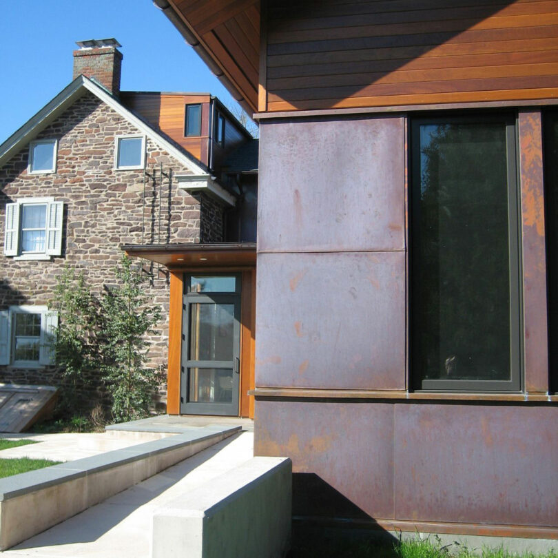 A modern extension with rust-colored metal panels by Barry Ginder adjoins a traditional stone house with white-trimmed windows under a clear blue sky.