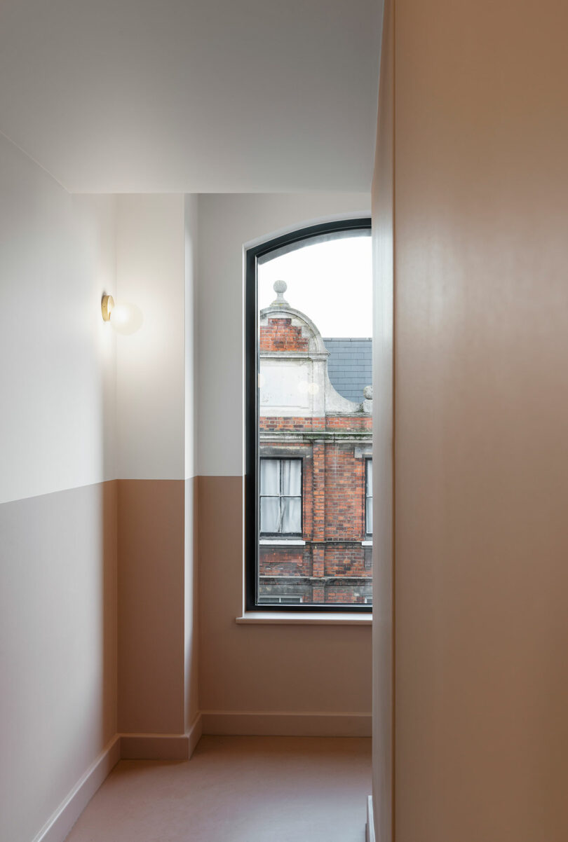 A minimalist hallway with two-tone walls, a round wall sconce, and an arched window overlooking an ornate brick building.