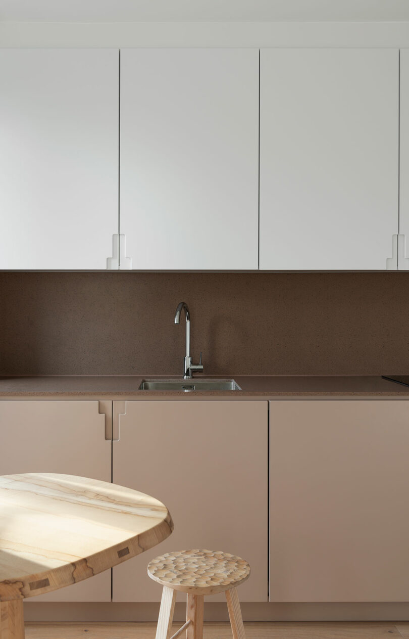 Minimalist kitchen with white upper cabinets, beige lower cabinets, brown backsplash, a metal sink with faucet, wooden table, and a wooden stool in the foreground.