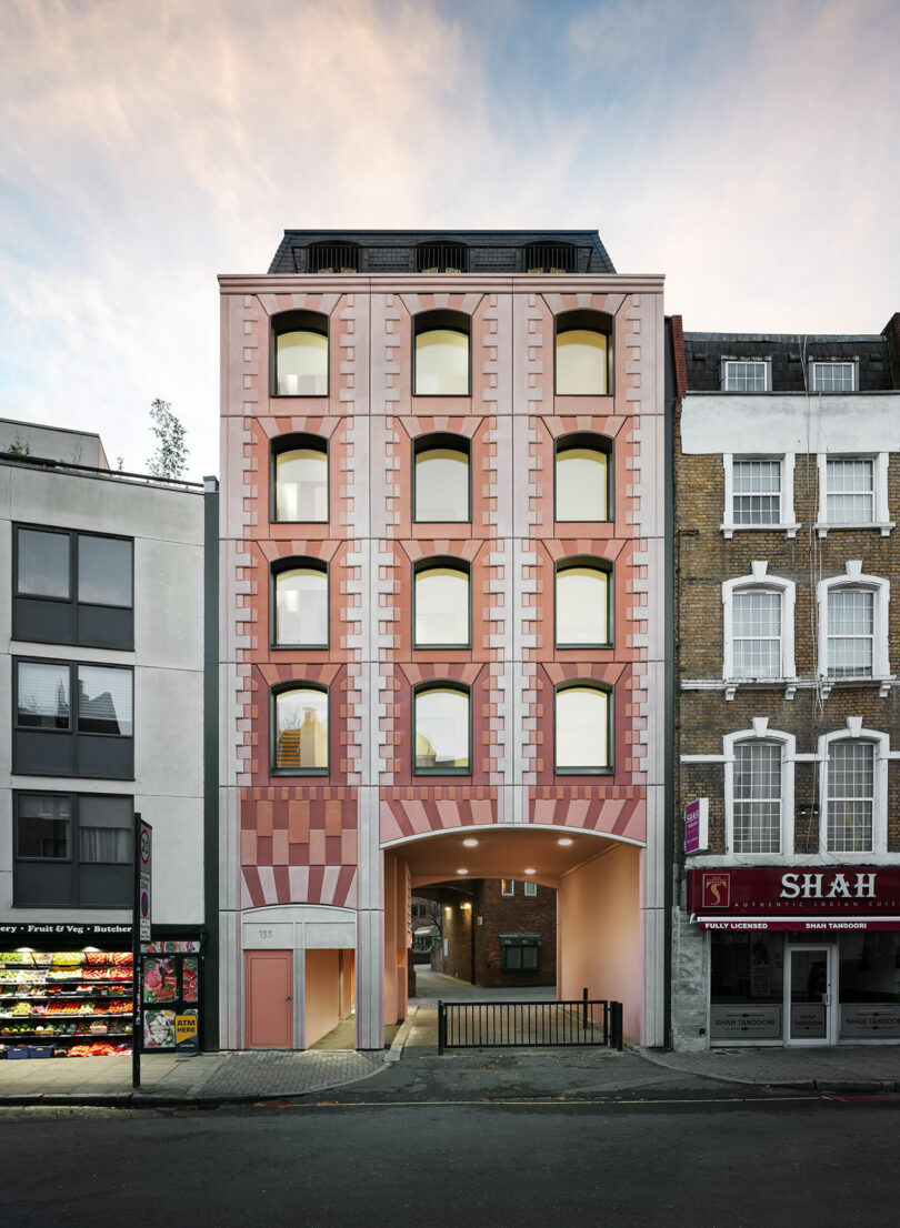 A modern pink and beige mid-rise building with arched windows stands between a grocery store and a restaurant on a city street.