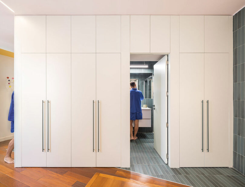 A person in a blue robe stands at a bathroom sink, visible through a doorway set within a wall of white storage cabinets.
