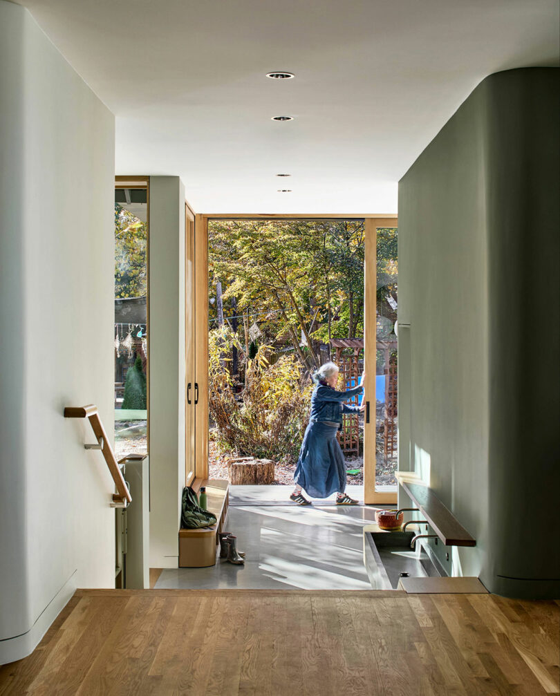 A person sweeps leaves outside a glass door, viewed from inside a modern home with wood flooring and minimalist decor.