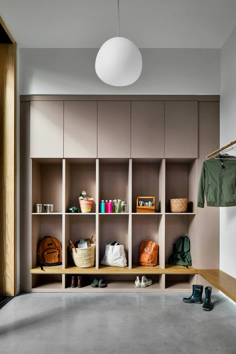 A modern mudroom with built-in cubbies holding bags, baskets, shoes, and various small items; a coat rack with a green jacket and a bench are also visible.