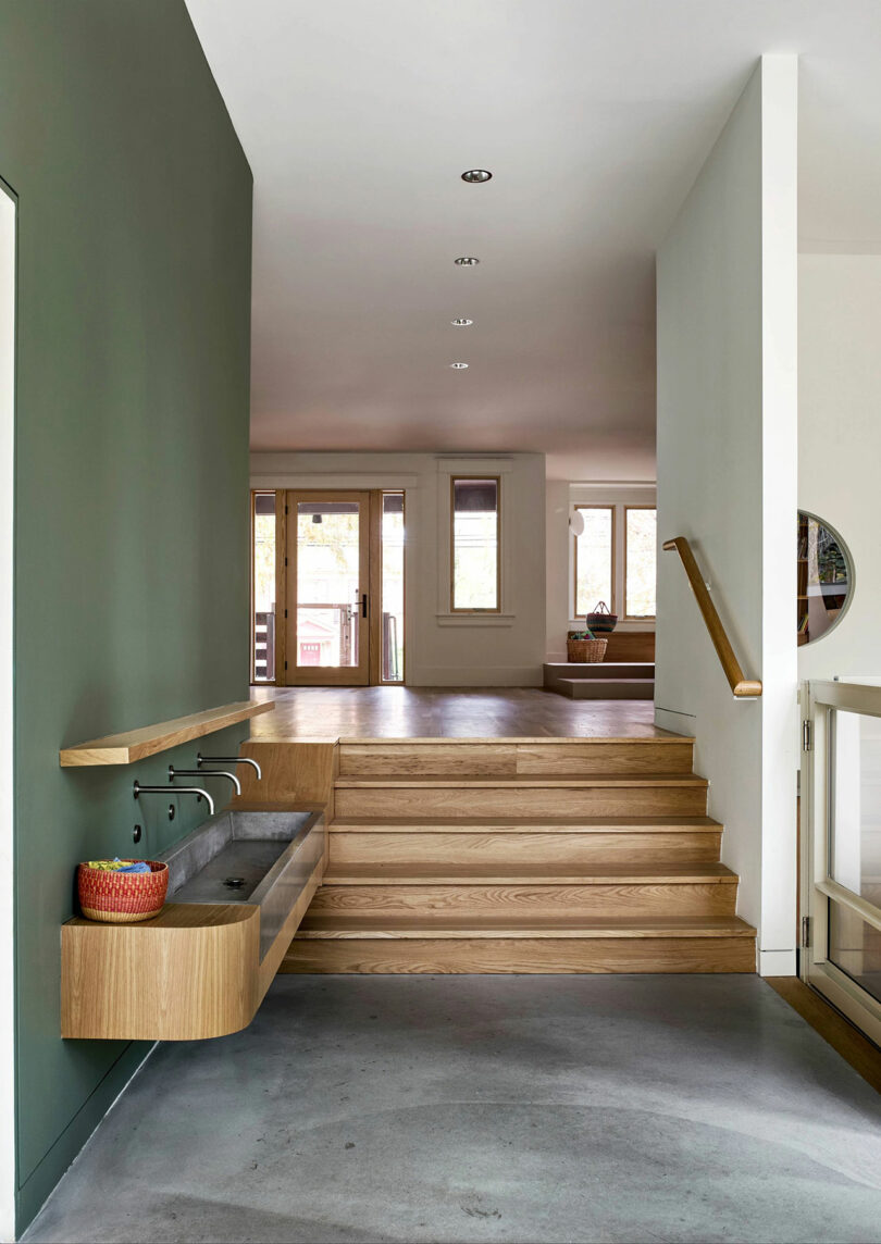 A modern interior with wooden steps, a floating concrete sink with a green wall, and doors leading to a bright room in the background.