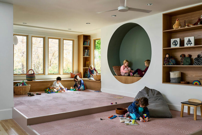 Children play with toys in a modern playroom featuring shelves, a large window, and a circular nook where two children sit and talk.