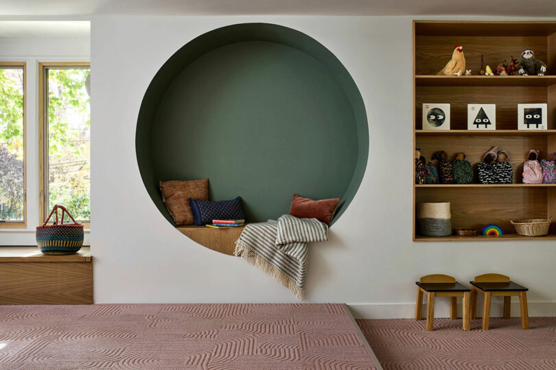 Room with a large circular wall nook containing cushions and a blanket, adjacent to shelves with toys and baskets, and two small stools on a patterned floor.