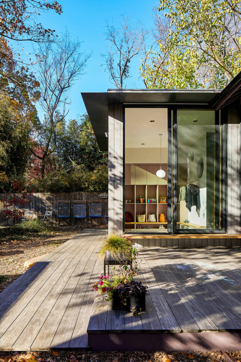 A modern house with large glass windows opens onto a wooden deck. Potted plants sit on the deck, and a shelving unit is visible inside the house. Trees surround the area.