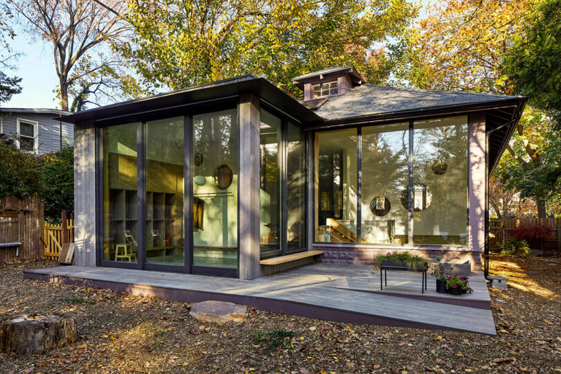 Modern house with large floor-to-ceiling glass windows and a small raised deck, surrounded by trees with autumn foliage.