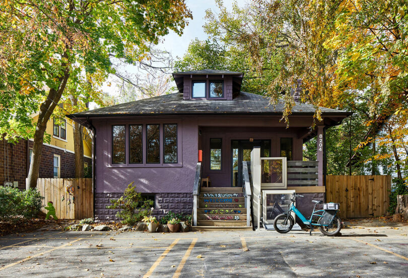 A purple single-story house with large windows, a bike parked by the porch steps, and autumn trees surrounding the building.