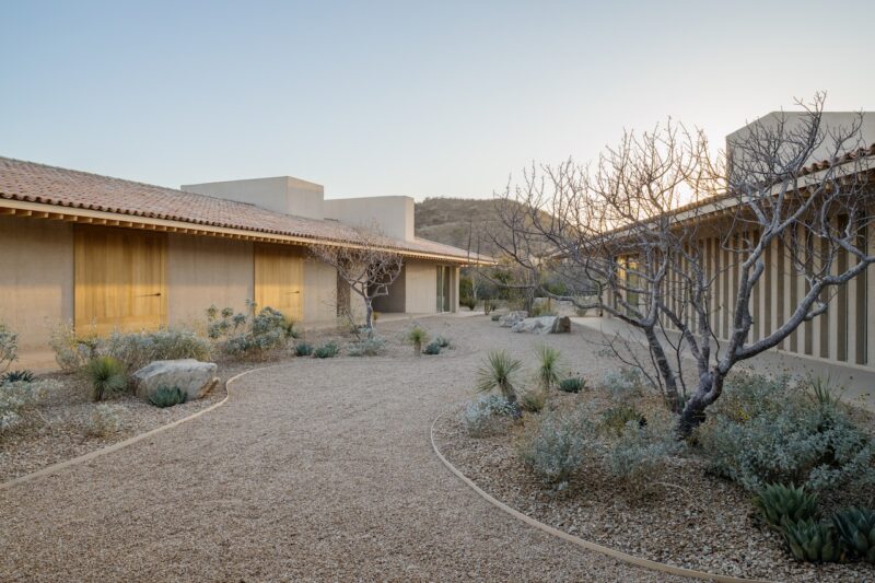 A gravel courtyard with desert plants and leafless trees is surrounded by beige, flat-roofed buildings under a clear sky at sunset.