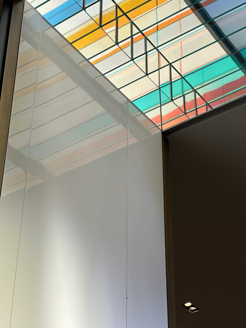 Glass wall reflecting a ceiling with colorful striped panels and a portion of a dark interior wall with recessed lights, creating an atmosphere perfect for Friday 5 gatherings.
