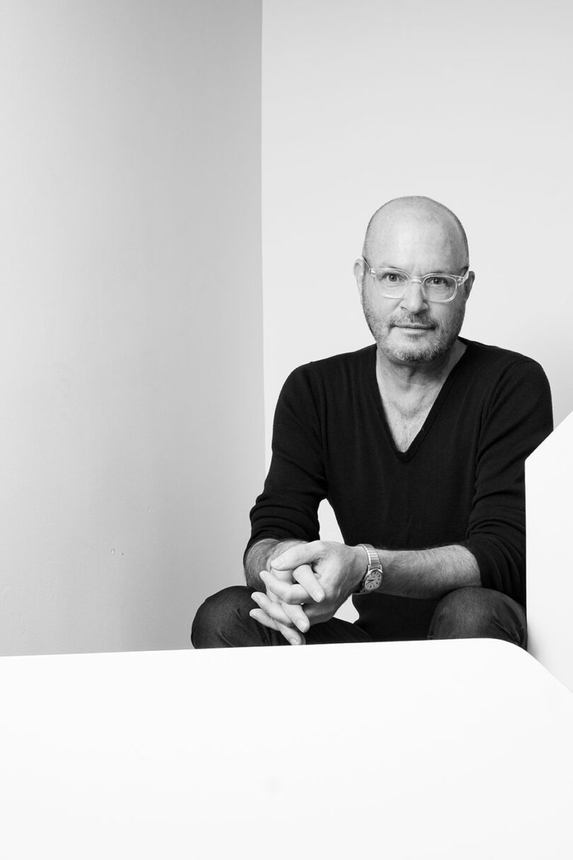A man with glasses and a beard sits against a plain background, wearing a dark V-neck sweater and watch, looking at the camera with hands clasped—capturing a calm Friday 5 mood in this black and white photo.