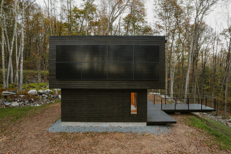 Modern two-story black cabin with large solar panels on the upper facade, surrounded by trees and featuring a small deck on the right side.