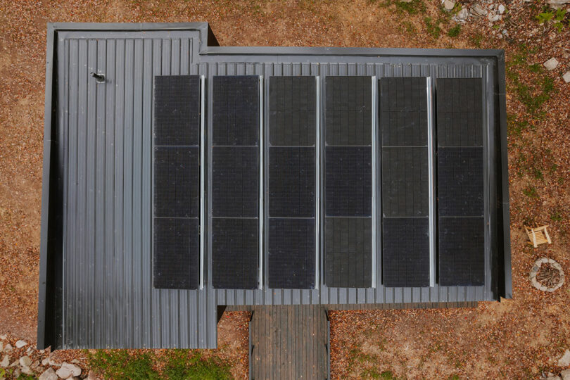 Aerial view of a gray metal roof with eight rectangular solar panels arranged in two rows, surrounded by dry ground and a small fire pit on the right.