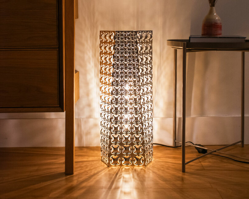 A decorative floor lamp with a geometric metal shade emits warm light, casting patterned shadows on the wall and floor; a wooden cabinet and a small table are nearby.