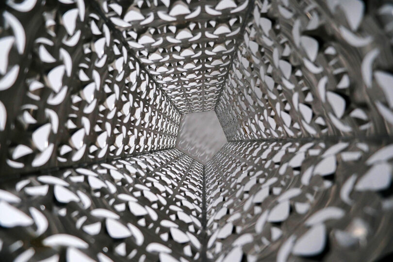 View looking upward through a metallic, geometric structure with intricate cut-out patterns and a pentagonal opening at the top.