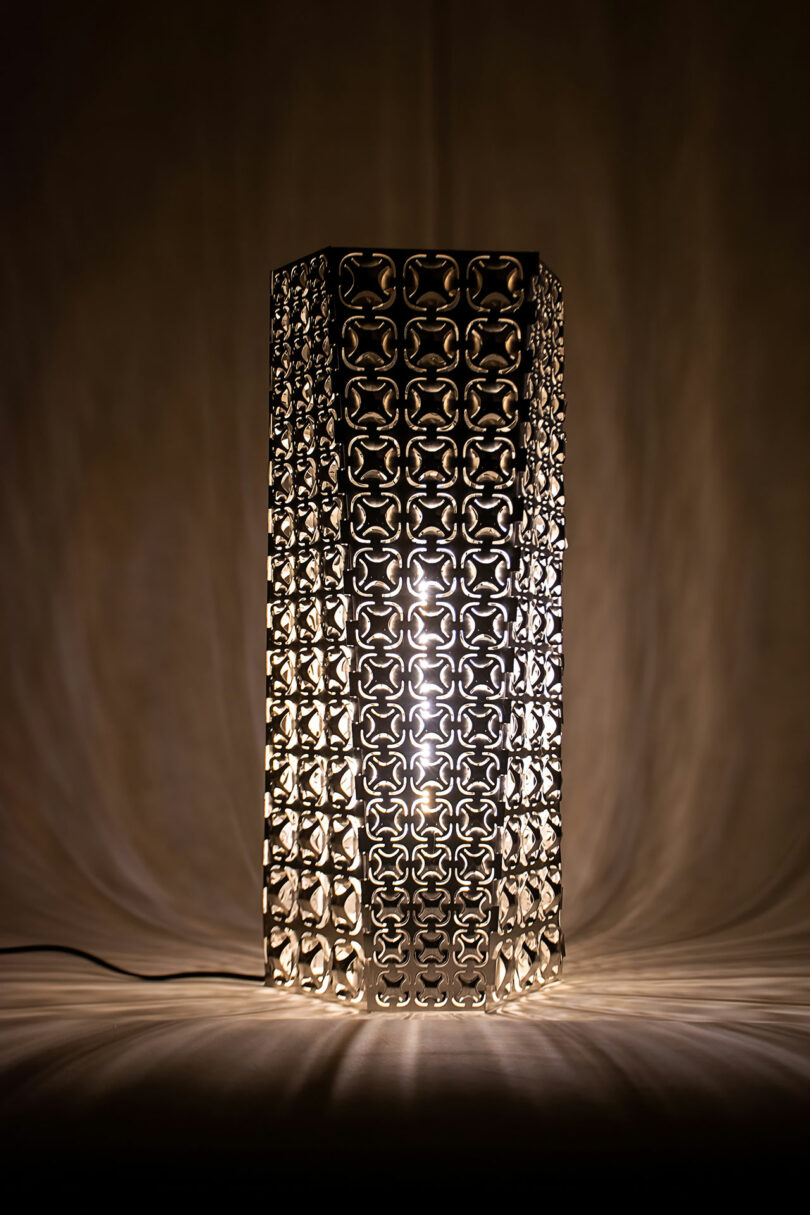 A geometric metal table lamp with a repeating pattern projects intricate shadows on a neutral background as it emits a warm light.