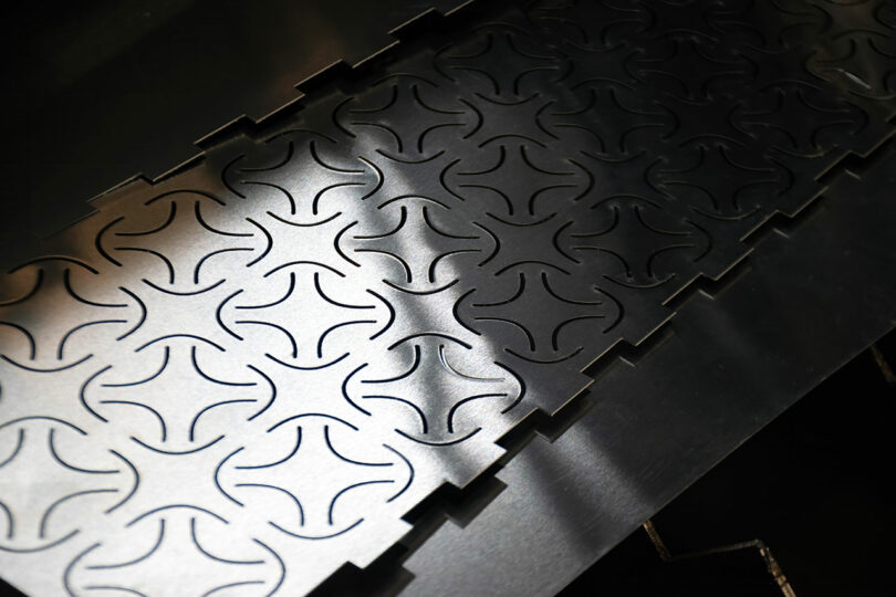 Sheet of metal with a repeating geometric pattern of interlocking curved lines cut into the surface, shown under direct lighting.