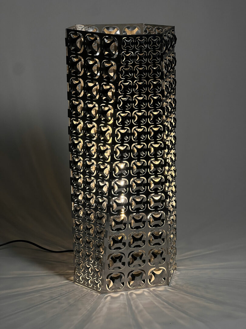 A geometric metal table lamp with intricate cutout patterns emits soft light, creating decorative shadows on a gray surface. The lamp is plugged in with a visible black cord.