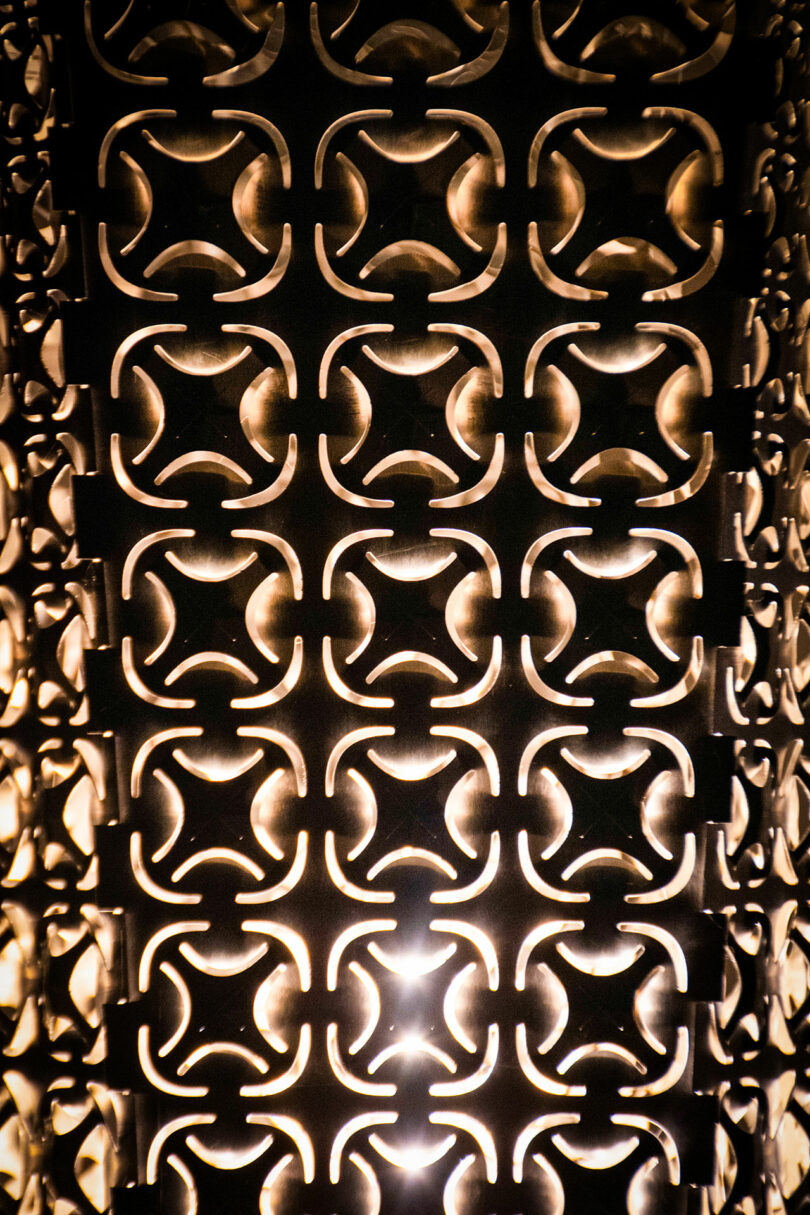 Close-up of an illuminated decorative metal panel with a repeating geometric cutout pattern, casting intricate shadows and light patterns.
