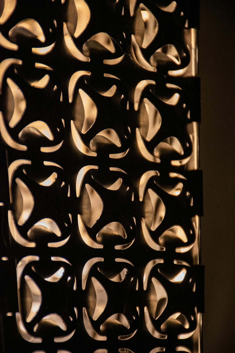 A close-up of a decorative panel with repeating abstract cut-out patterns, illuminated from behind by warm light.