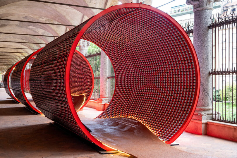 Large cylindrical red tunnel art installation with an open walkway underneath, set in a covered outdoor corridor with columns and iron fencing.